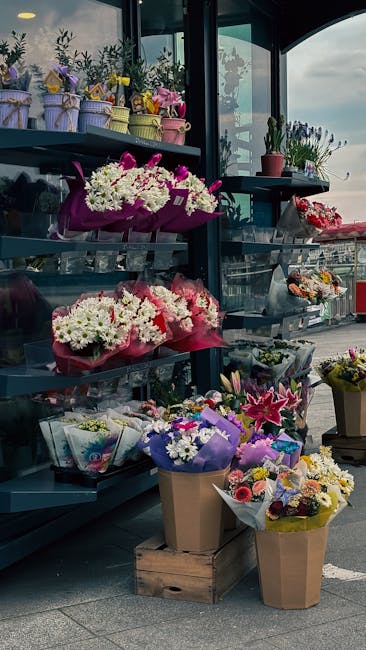A display of various floral arrangements and bouquets outside a shop, featuring vibrant pink, white, purple, and yellow flowers, including chrysanthemums, lilies, and roses. The flowers are wrapped in