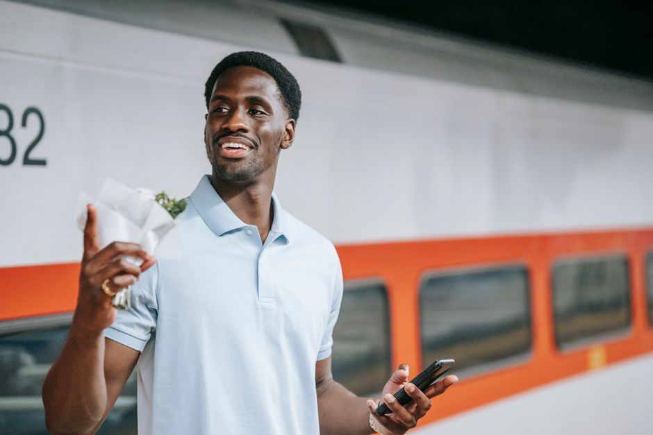 A smiling young man with dark skin and short black hair is holding a small bouquet of fresh flowers wrapped in white paper in his right hand and a smartphone in his left hand. He is standing on a trai